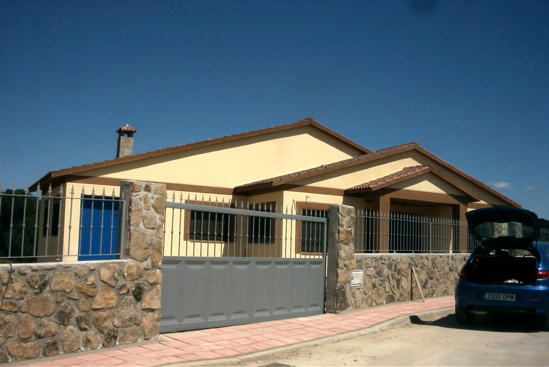 Casa de una sola planta color canela con muro de piedra y portón gris; coche azul aparcado cerca. Cielo despejado.