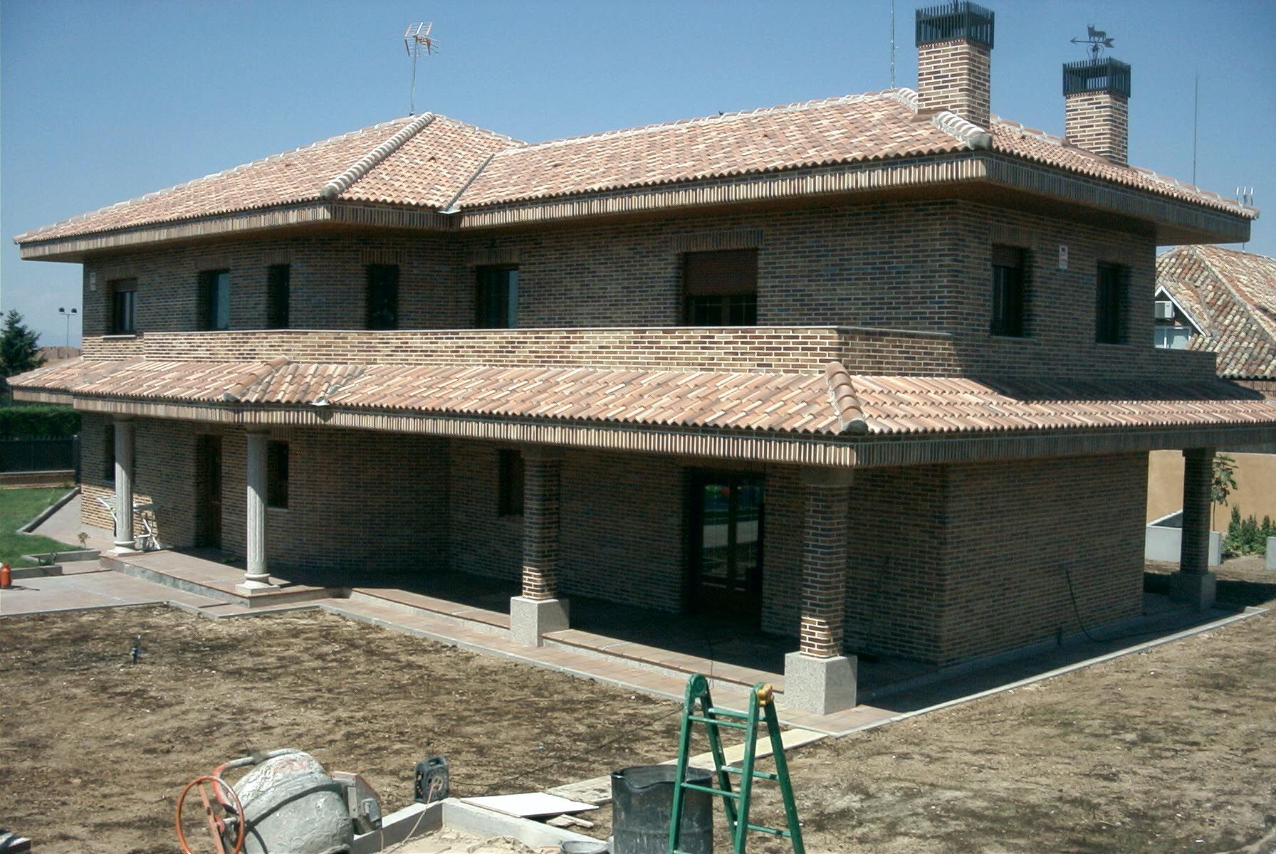 Casa de ladrillo de dos plantas con techo de teja, columnas y dos chimeneas. En una obra en construcción.