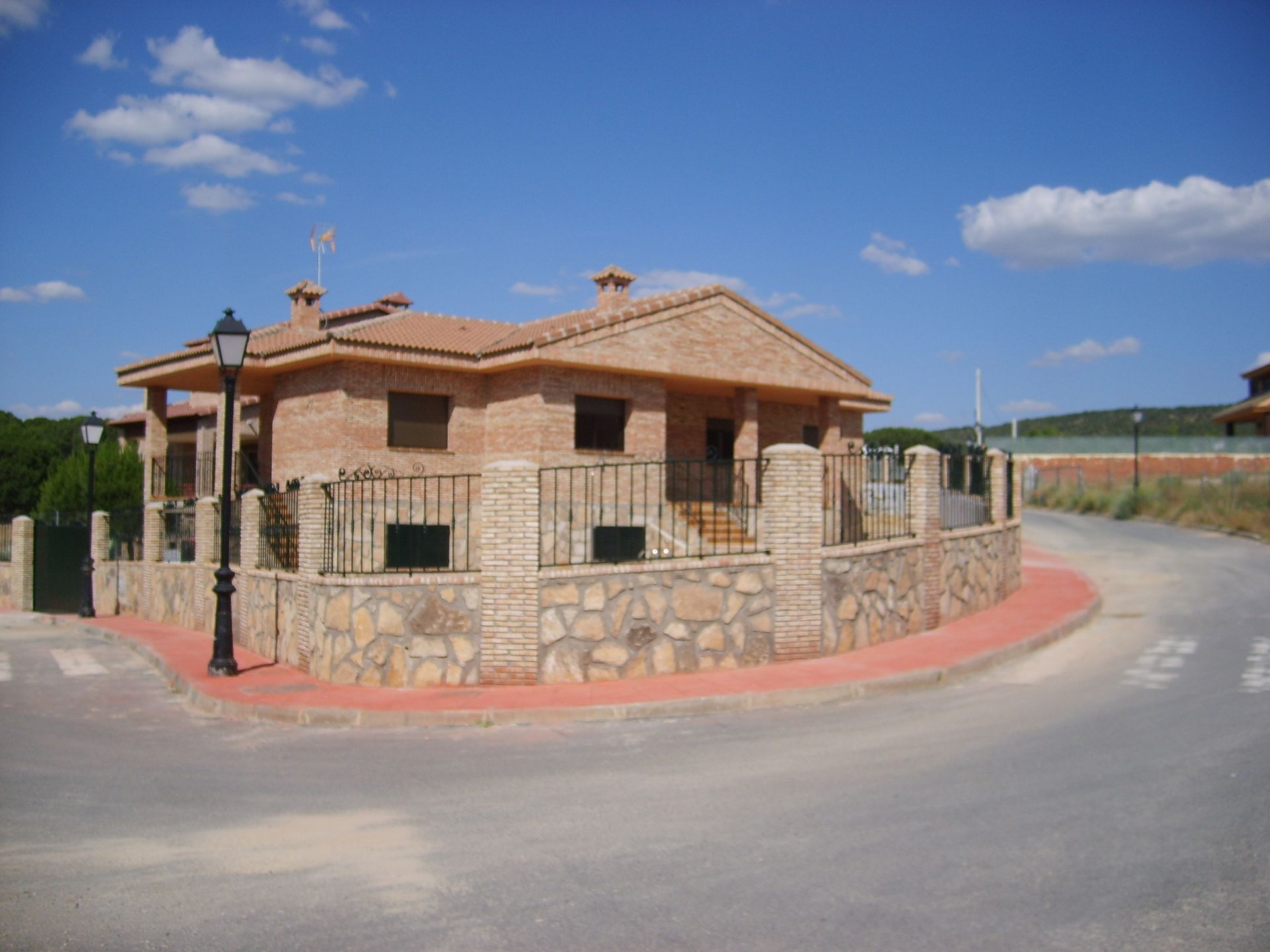 Casa de ladrillo en una esquina de la calle con muro de piedra y valla, cielo azul con nubes.