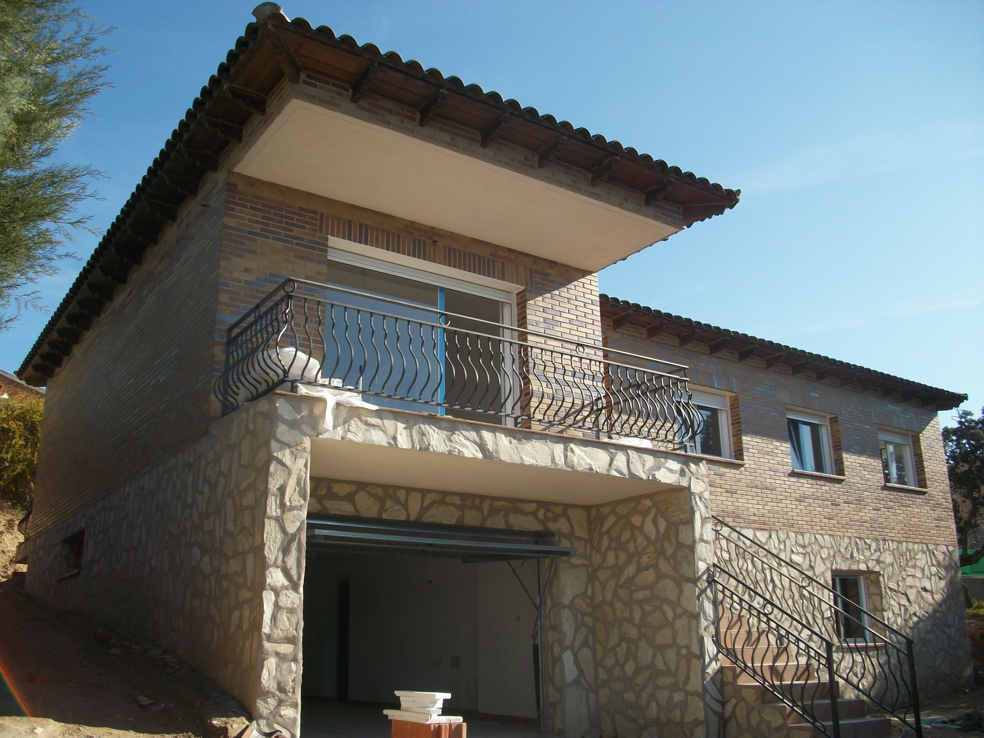 Casa de dos plantas con exterior de piedra y ladrillo; balcón y garaje bajo el balcón. Cielo azul.