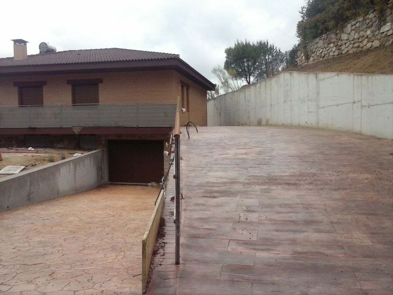 El camino de entrada marrón asciende hacia una casa con garaje, flanqueado por muros de contención de hormigón.