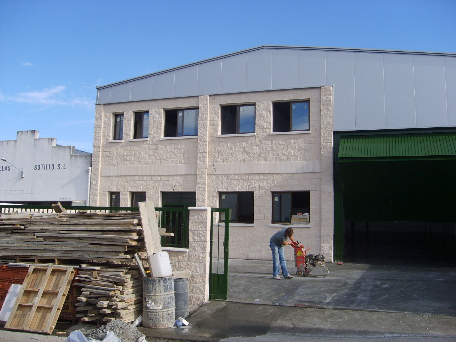 Un edificio de dos plantas con un hombre usando una herramienta eléctrica en la entrada. Hay palets de madera a la izquierda.