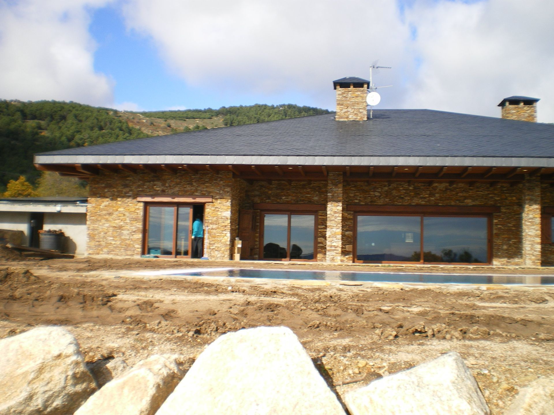 Casa de piedra con grandes ventanales, tejado de pizarra y piscina en construcción.