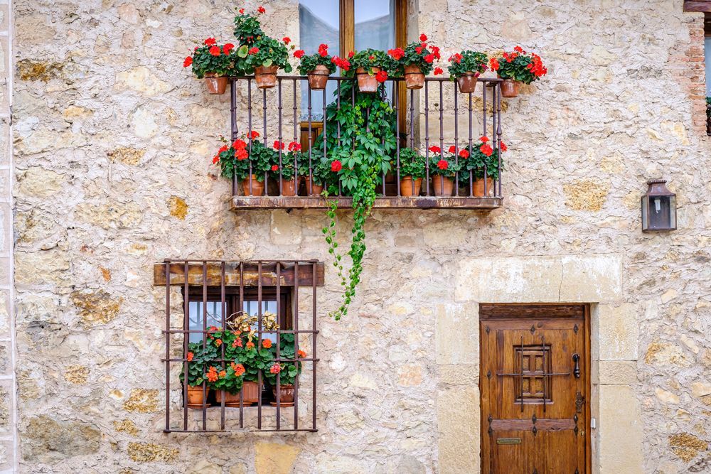 Fachada de edificio de piedra con puerta de madera, ventana enrejada y balcón rebosante de flores rojas.
