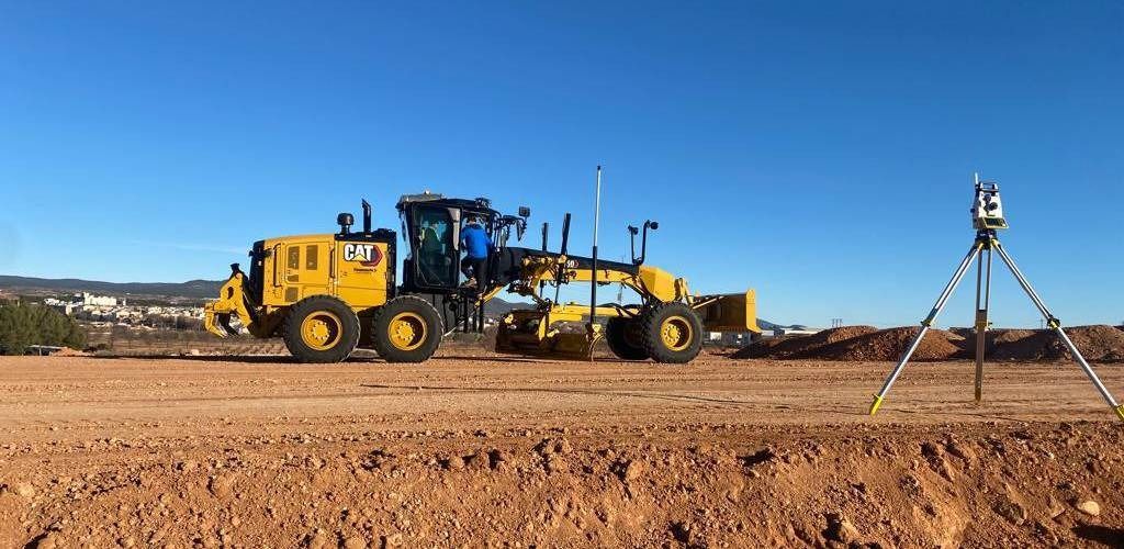 Un tractor amarillo circula por un campo de tierra al lado de un trípode.
