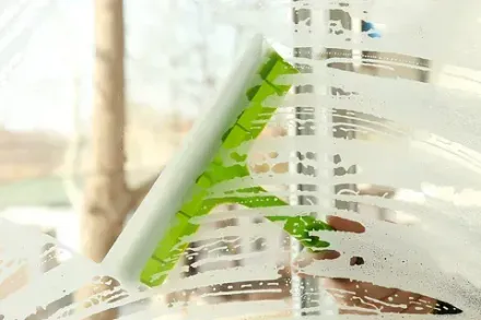 Una persona que usa una escobilla de goma verde para limpiar una ventana enjabonada, con un árbol borroso visible afuera.