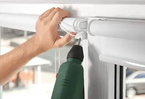 Persona que utiliza un taladro eléctrico para instalar una persiana blanca.