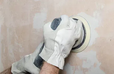 Una persona que usa guantes de trabajo lija un parche en una pared de color marrón claro con una lijadora circular.