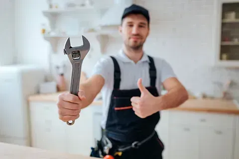 Fontanero con mono sosteniendo una llave inglesa y haciendo un gesto de aprobación con el pulgar en una cocina.