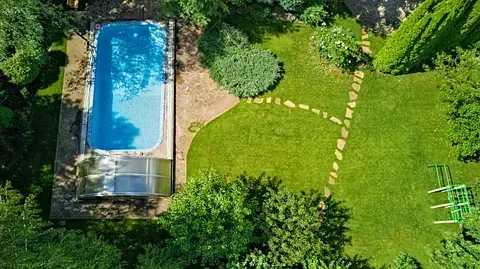 Vista aérea de una piscina rectangular rodeada de exuberante césped verde, árboles y un camino de piedra.