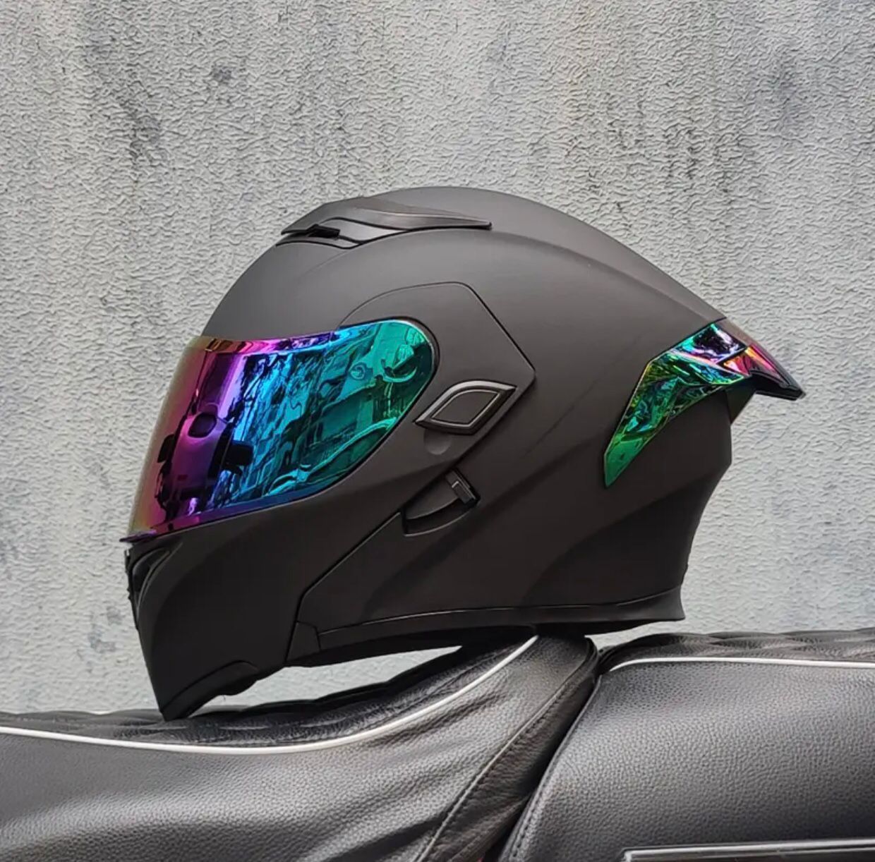 Casco de motocicleta negro mate con visera iridiscente, apoyado sobre un asiento negro contra una pared gris claro.