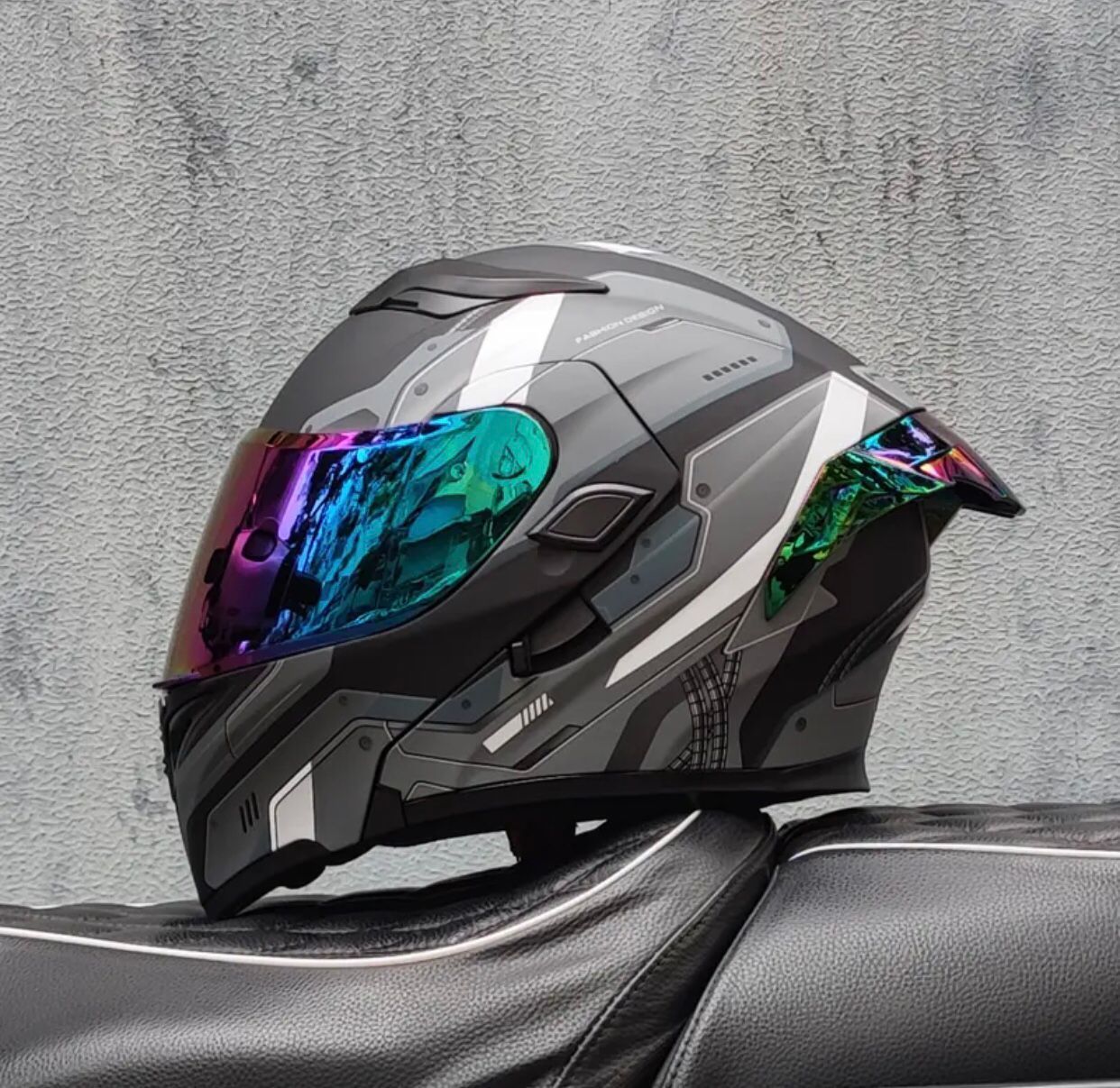 Un casco de motocicleta gris y plateado con una visera colorida se encuentra sobre un asiento negro.