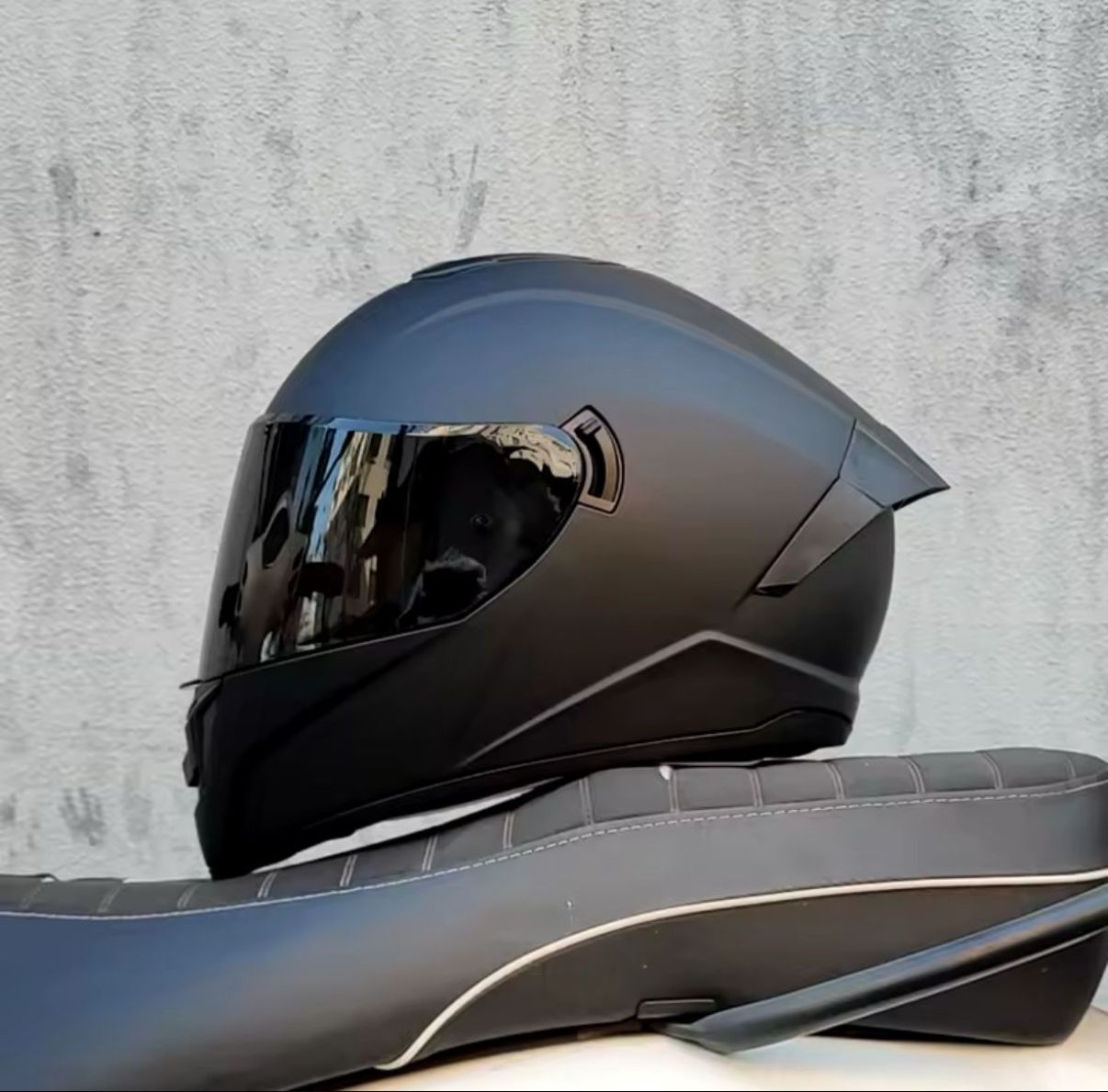 Casco de motocicleta negro con visera tintada sobre un asiento negro contra un fondo gris.