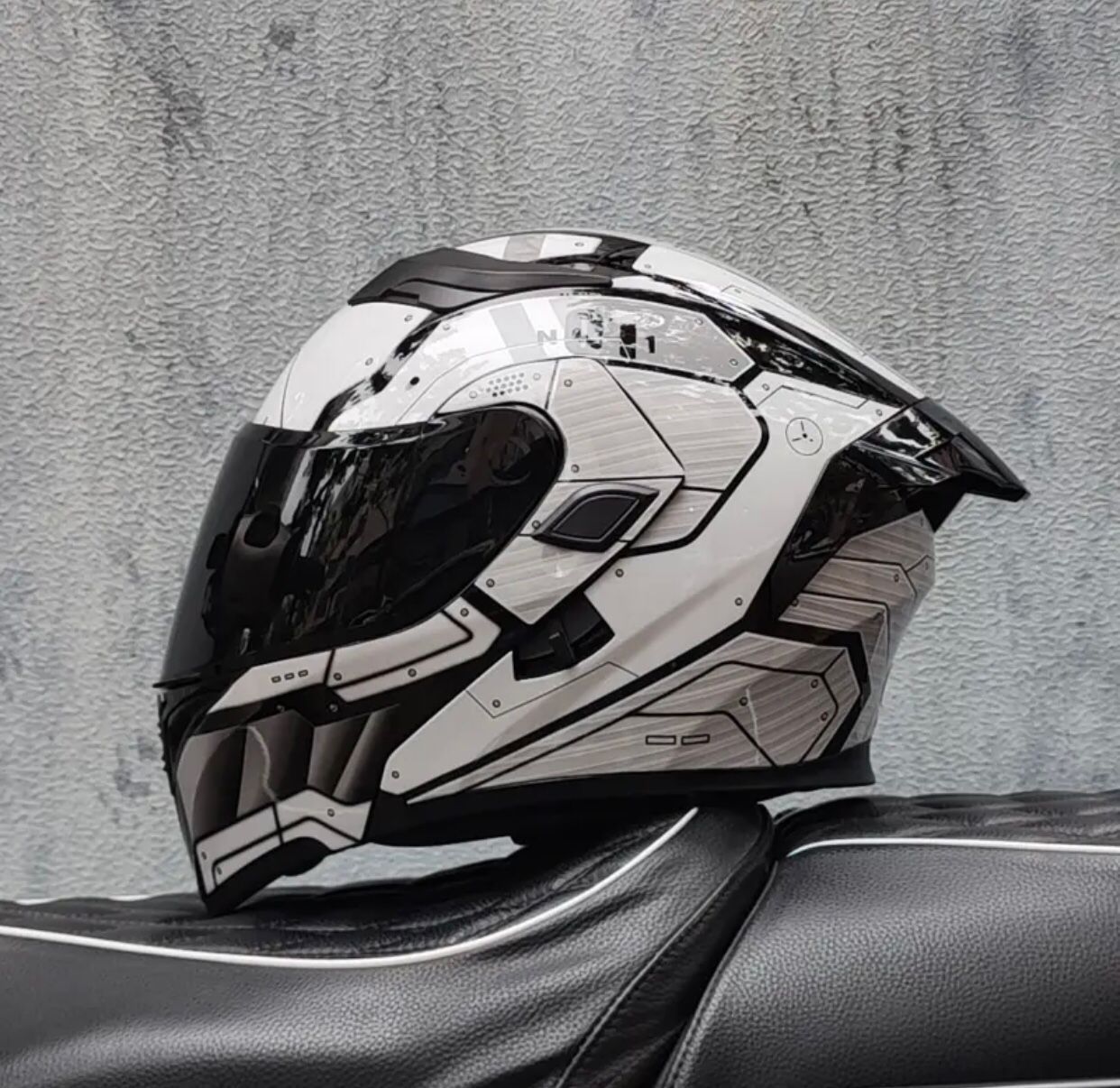 Casco de motocicleta blanco y gris con diseños geométricos, sobre un asiento negro. Visera negra cerrada.