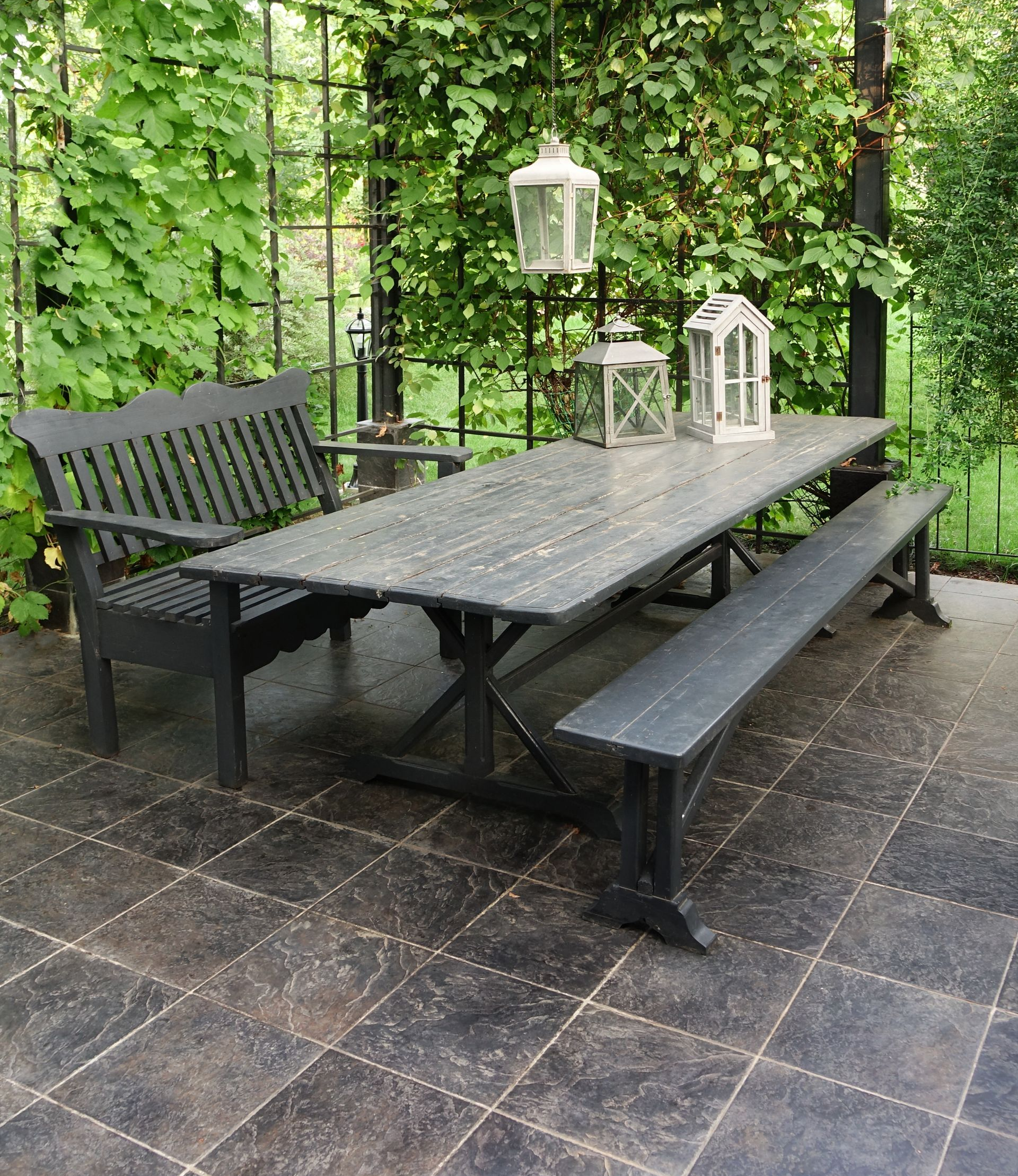 Ensemble de repas en plein air sur une terrasse carrelée, comprenant une table gris foncé, un banc et un fauteuil avec des lanternes. Végétation luxuriante en arrière-plan.