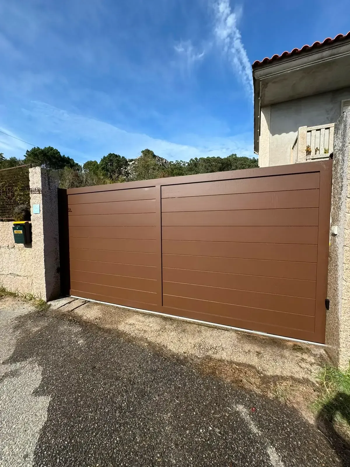 Una puerta metálica marrón junto a la pared de estuco de una casa bajo un cielo azul, con un camino de entrada pavimentado al frente.