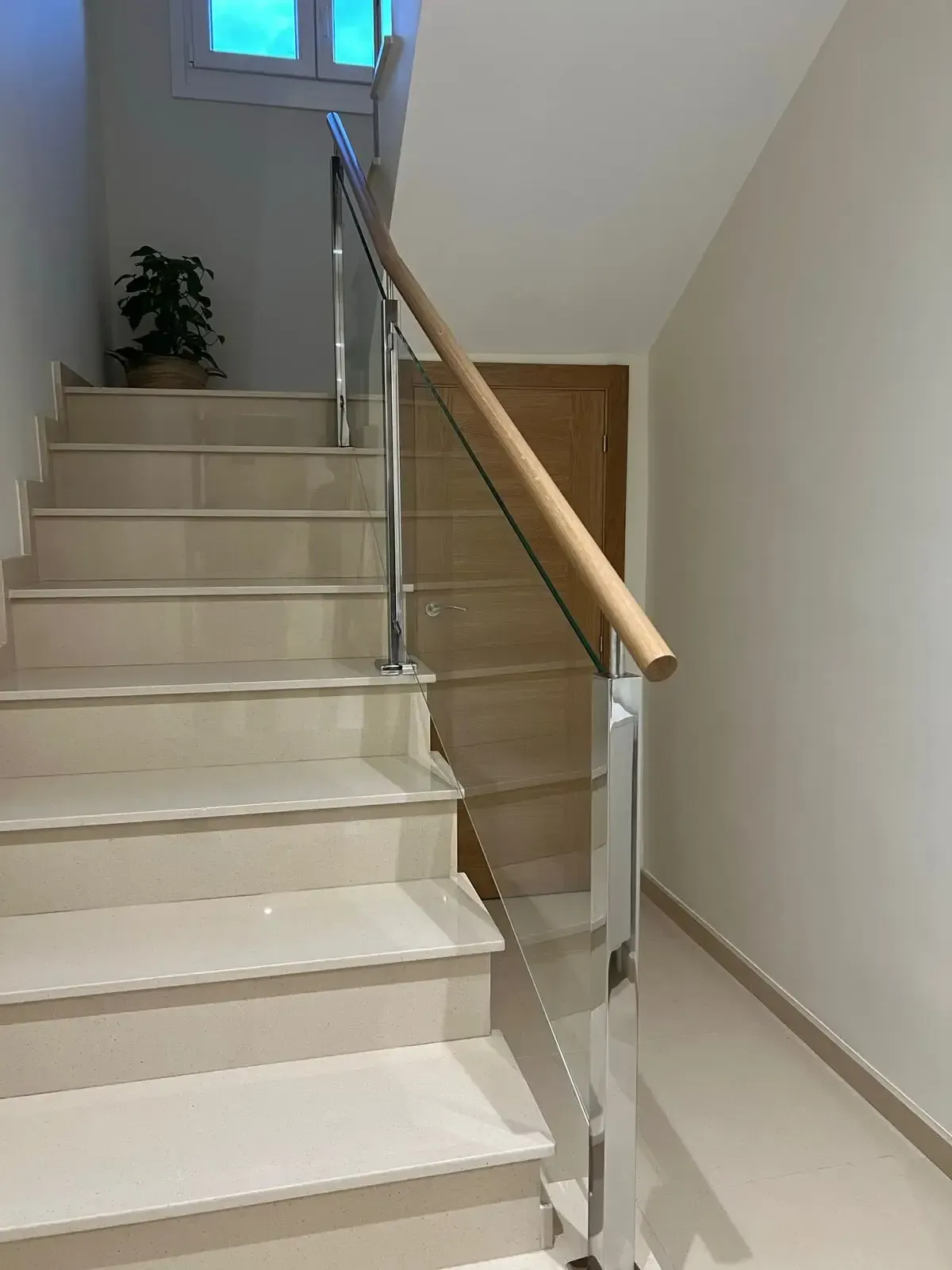 Escalera interior con escalones color crema, barandilla de cristal y pasamanos de madera que conduce a una ventana luminosa.
