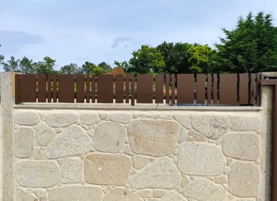 Muro de piedra con valla de madera marrón y árboles verdes detrás bajo un cielo azul
