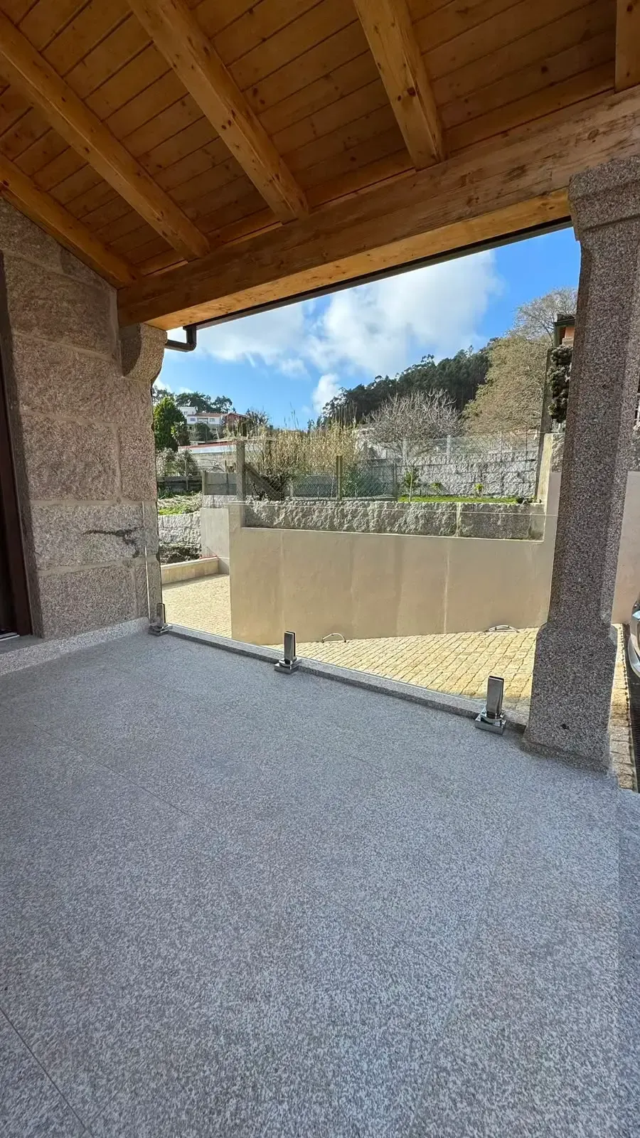 Patio cubierto de piedra con techo de madera, con vistas a un jardín vallado y árboles bajo un cielo azul.