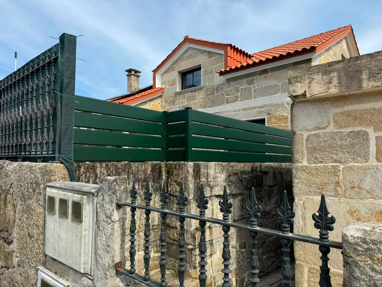 Muro de piedra y valla verde frente a una casa con tejas rojas bajo un cielo nublado.