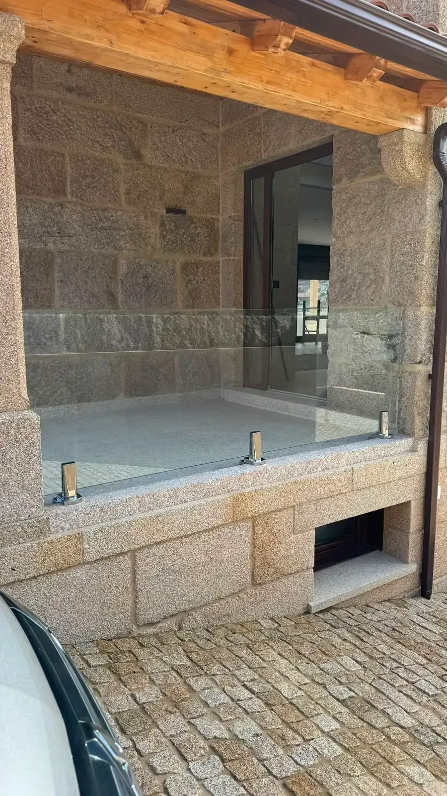 Porche de piedra con barandilla de cristal y patio empedrado junto a una puerta abierta.