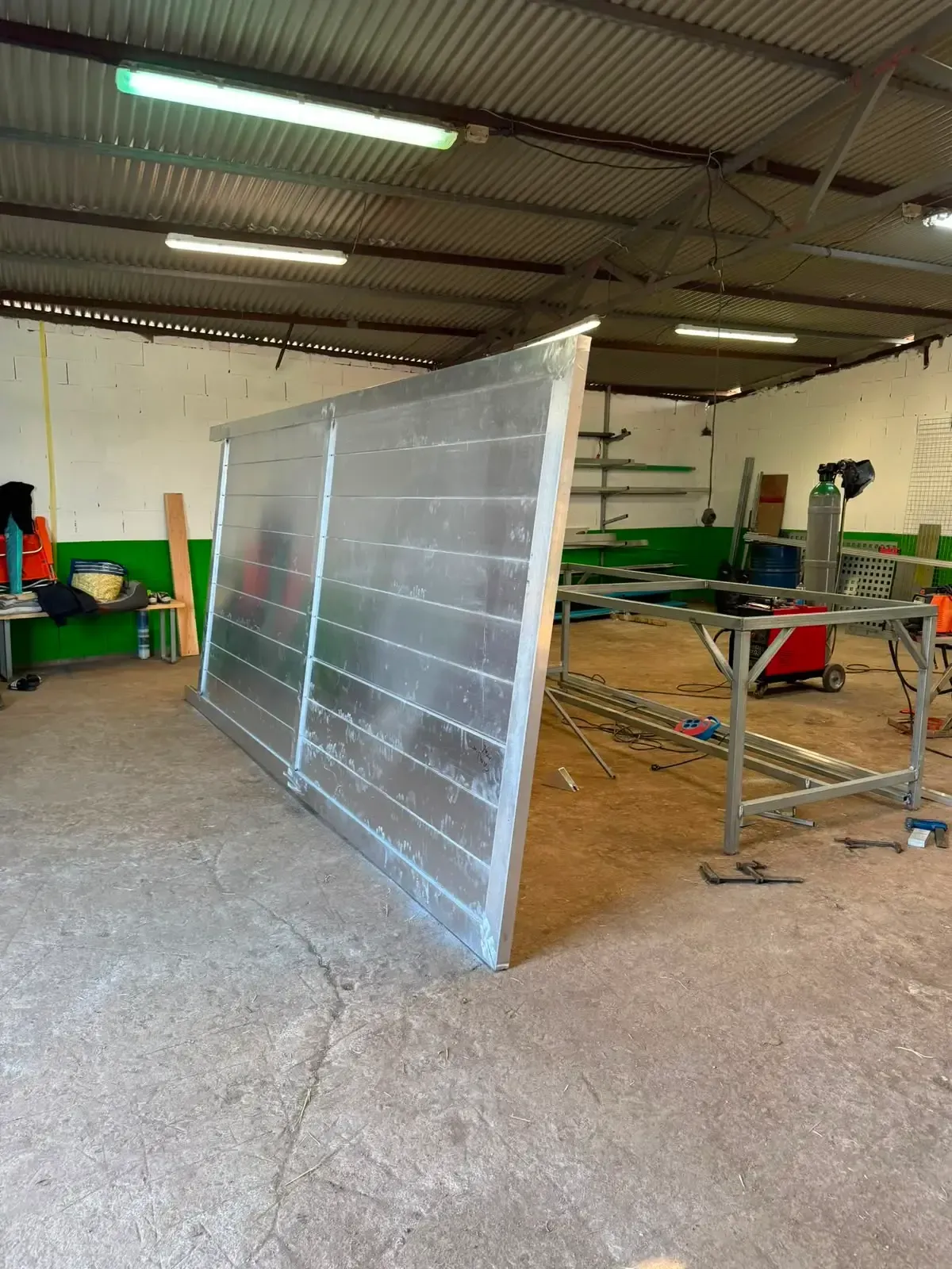 Gran panel metálico apoyado en un taller-almacén, con herramientas y equipos al fondo.