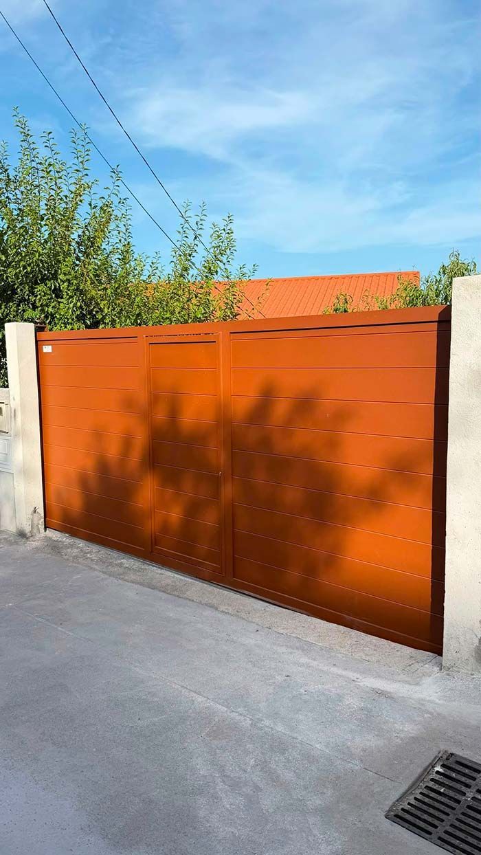 Portón metálico naranja junto a un muro de hormigón en una calle tranquila bajo un cielo azul