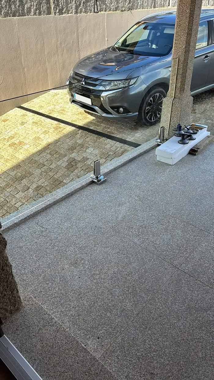 Un coche gris oscuro aparcado junto a un muro, cerca de un árbol, en un aparcamiento pavimentado