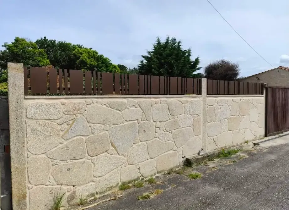 Muro de piedra con valla de madera marrón a lo largo de una calle residencial bajo un cielo nublado.