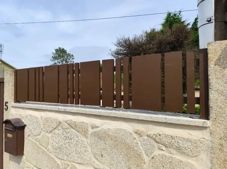 Una valla metálica marrón se alza sobre un muro de piedra clara frente a una casa, con un poste de servicios públicos a la derecha.