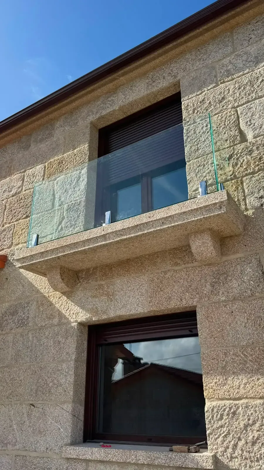 Exterior de una casa de piedra con dos ventanas y un pequeño balcón bajo un cielo azul.