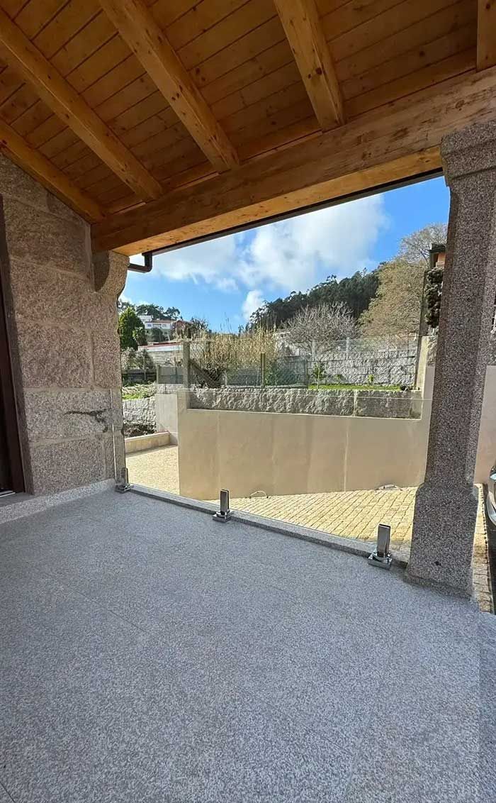 Patio de piedra cubierto con techo de madera, con vistas a un patio cercado y una ladera bajo un cielo azul