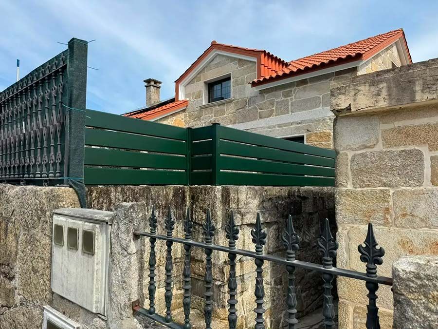 Casa de pedra con tellado de tella vermella e valado verde detrás dunha cancela metálica negra e dun muro de pedra.