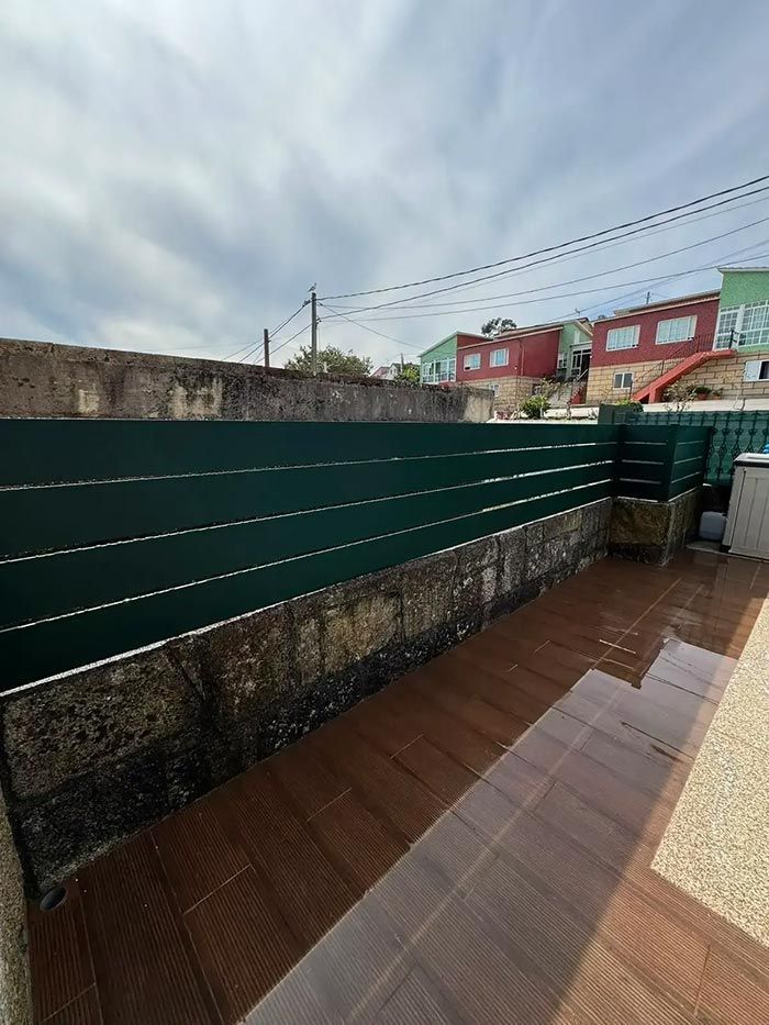 Balcón con paneis verdes de privacidade, chan de baldosa e vistas das casas e do ceo tras o muro