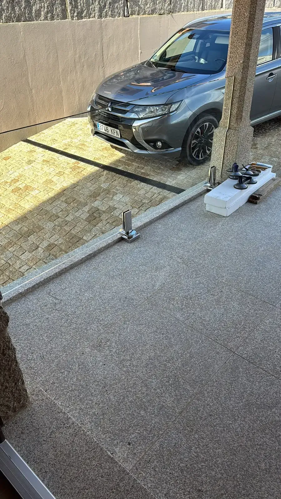 Un SUV gris estacionado junto a un muro de hormigón, visto desde un patio con un árbol y herramientas de limpieza en primer plano.