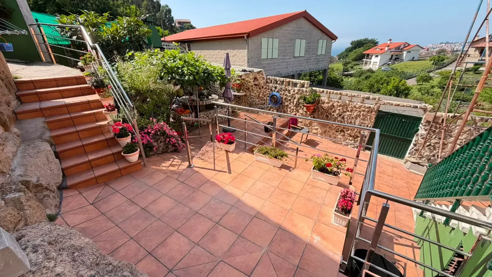 Patio ajardinado en terrazas con macetas de flores, escalones de piedra y vistas a las casas bajo un cielo azul brillante.