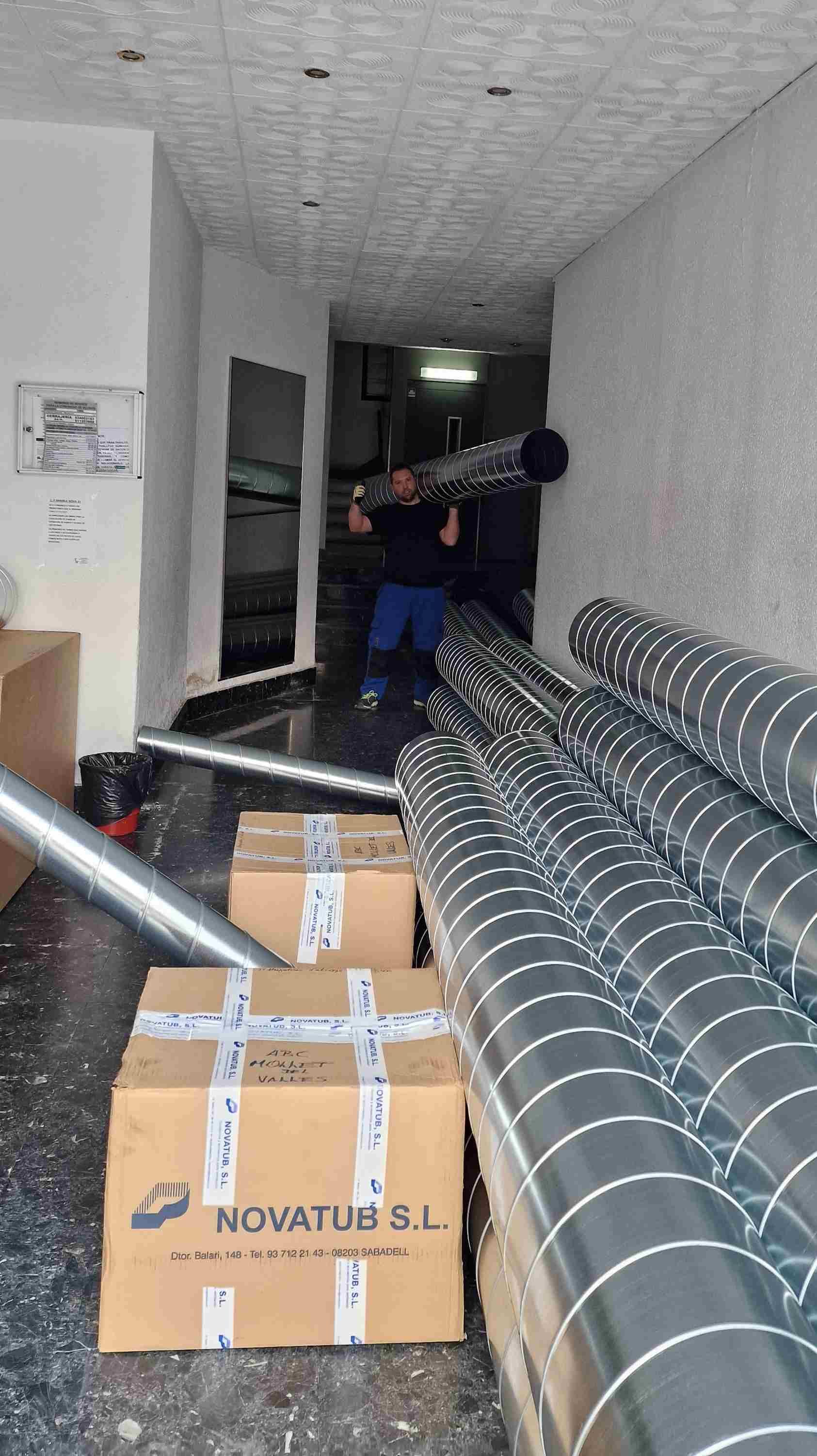 Hombre transportando conductos de metal por un pasillo desordenado, con cajas y más conductos en el suelo.