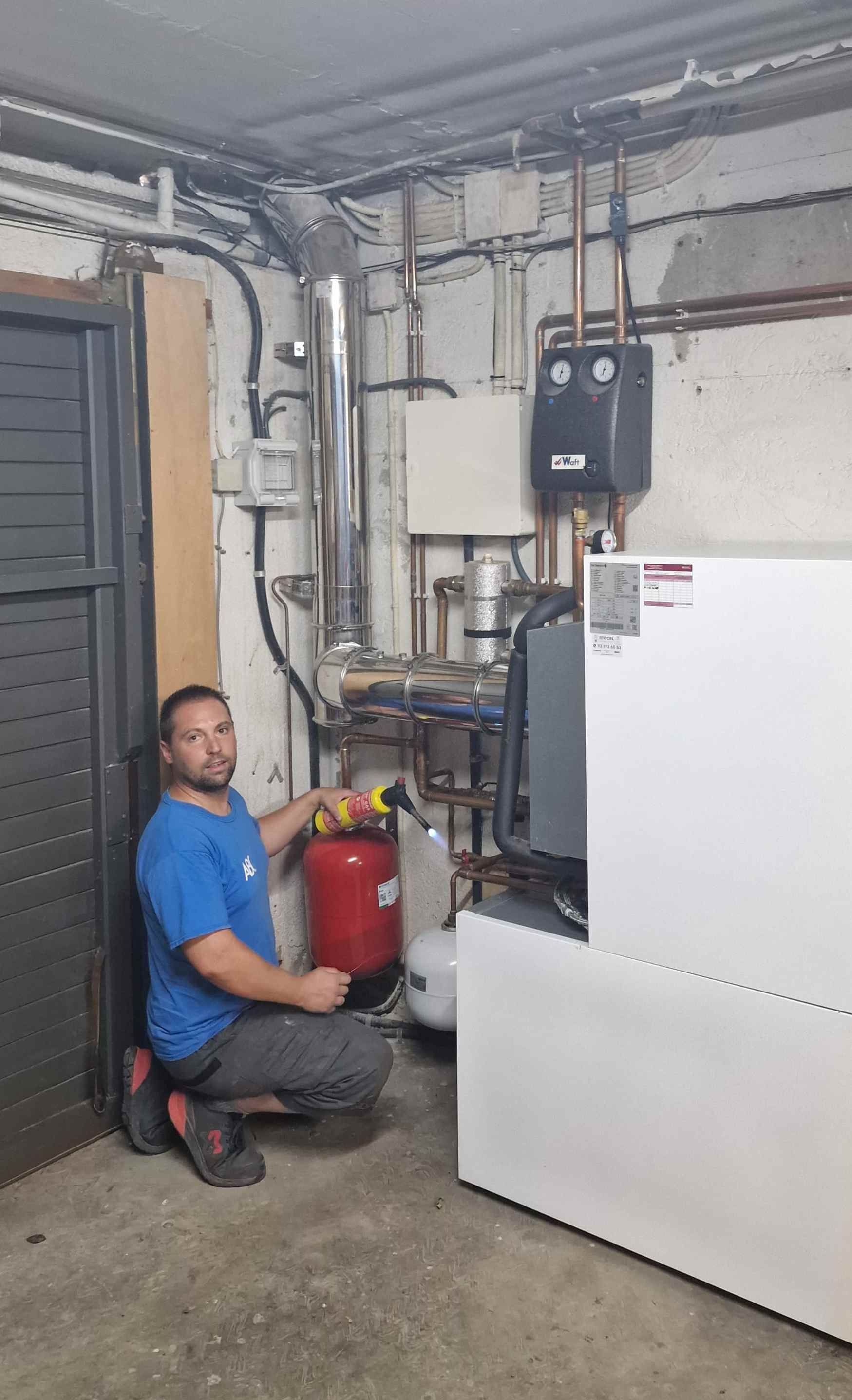 Un hombre de azul se arrodilla, sosteniendo un tanque rojo en un lavadero. Tuberías y maquinaria llenan las paredes.