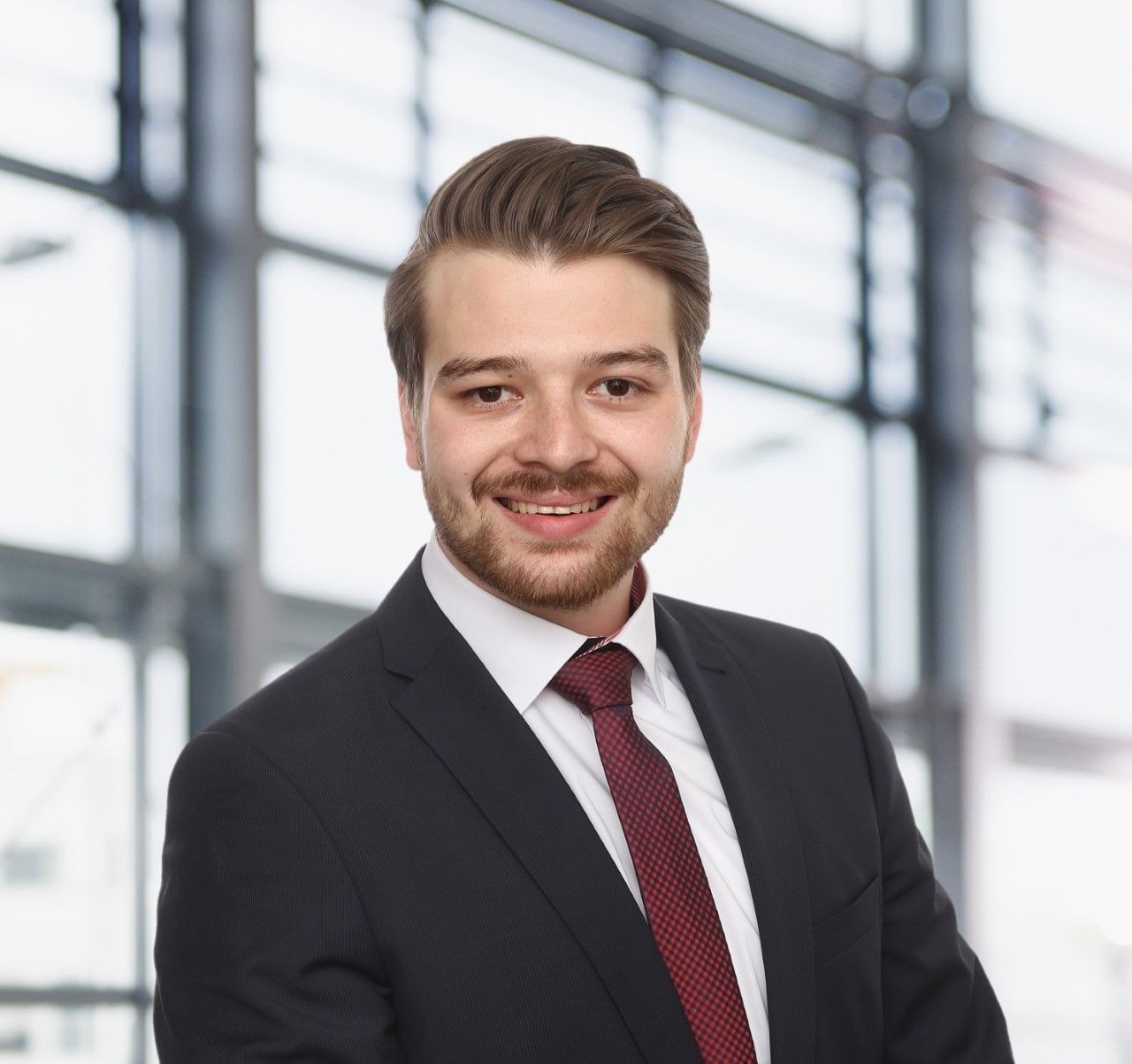 Ein lächelnder Mann in Anzug und Krawatte vor dem Hintergrund eines Bürofensters.