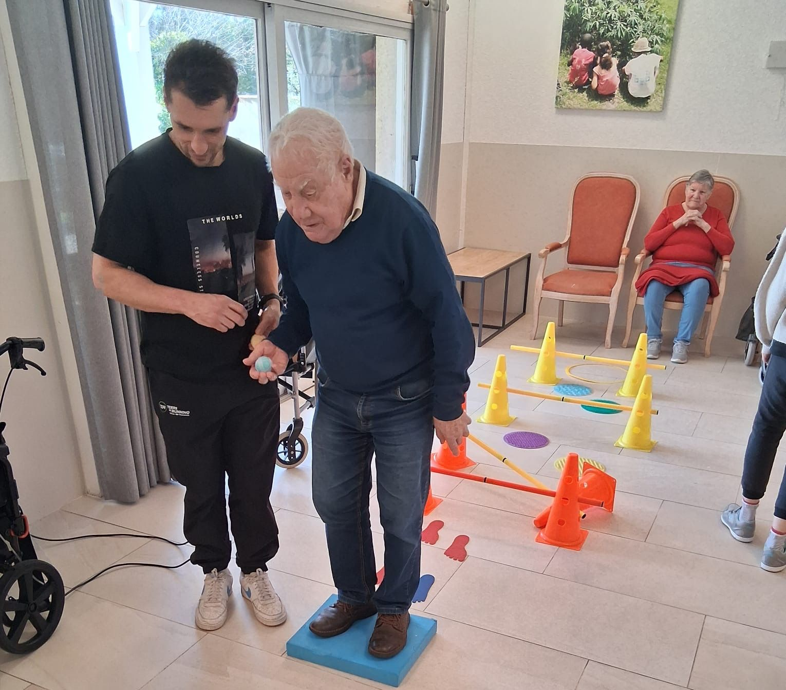 Dans une salle de rééducation, un homme âgé et un kinésithérapeute font un exercice de mobilité.