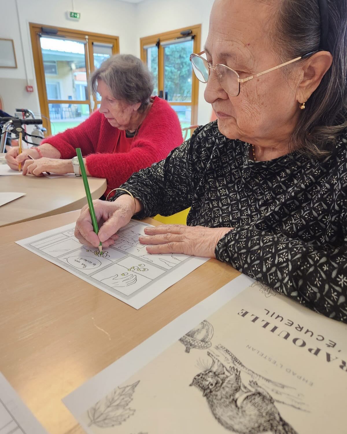 Des personnes colorient des feuilles de travail à une table de classe, une personne portant un pull noir au premier plan.