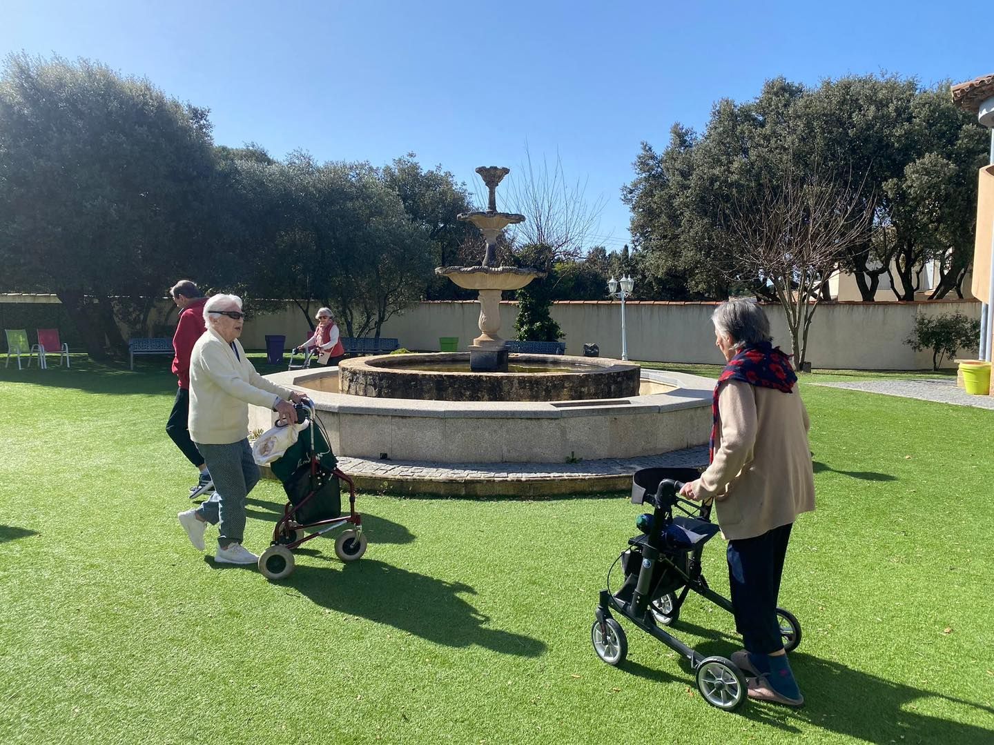 Deux personnes marchant avec des aides à la mobilité sur une pelouse autour d'une fontaine en pierre.