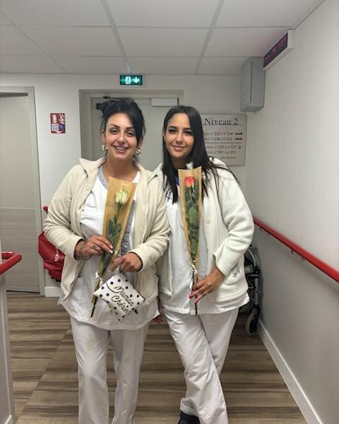 Deux personnes vêtues de blanc, tenant des fleurs, sourient à l'objectif dans un couloir.
