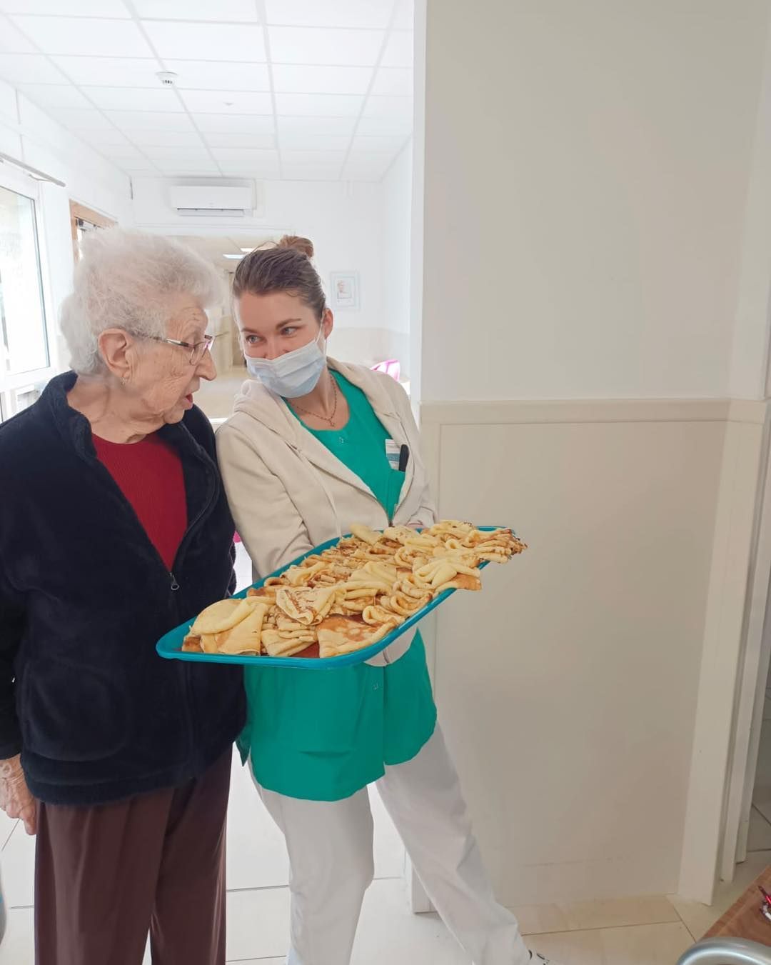 Une infirmière d'EHPAD tient un grand plateau de crêpes et discute avec une personne âgée.
