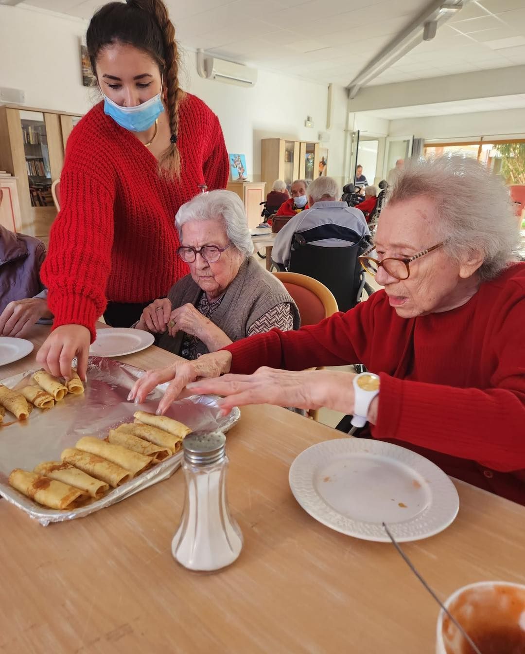 Une aide-soignante sert le repas à deux résidents attablés dans une salle commune lumineuse.