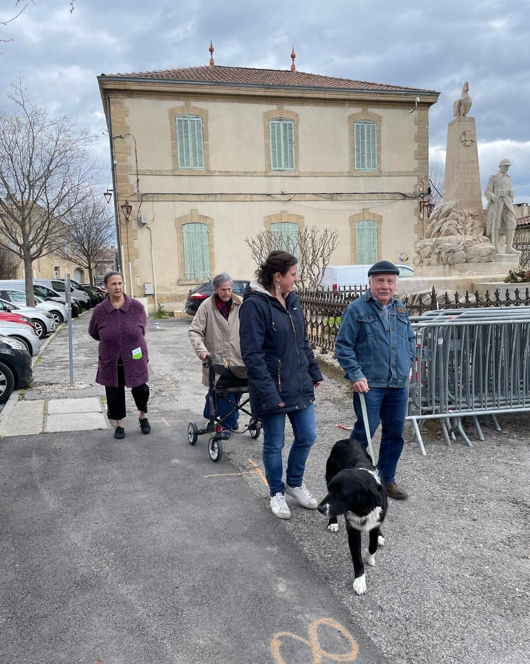 Des personnes promènent leur chien près d'un bâtiment historique et d'une fontaine par temps nuageux.