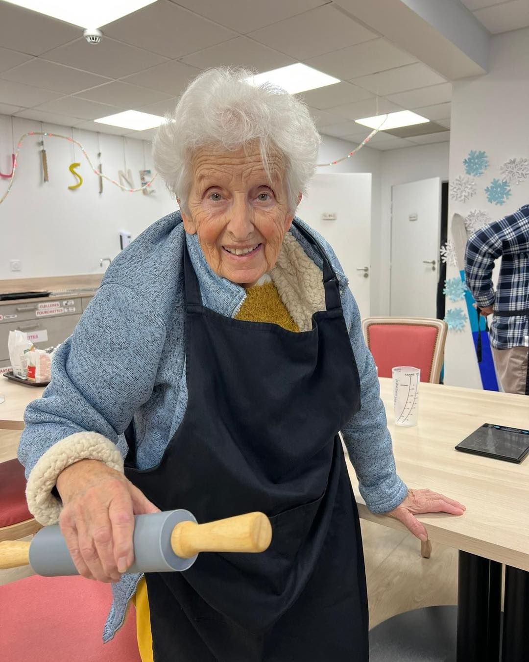 Femme souriante en pull bleu et tablier noir tenant un rouleau à pâtisserie dans une salle de classe ou un atelier.