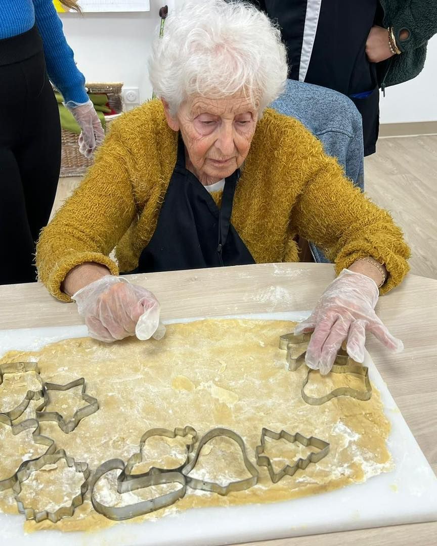 Une femme âgée vêtue d'un pull moutarde façonne de la pâte à biscuits à l'aide d'emporte-pièces, assise à une table.