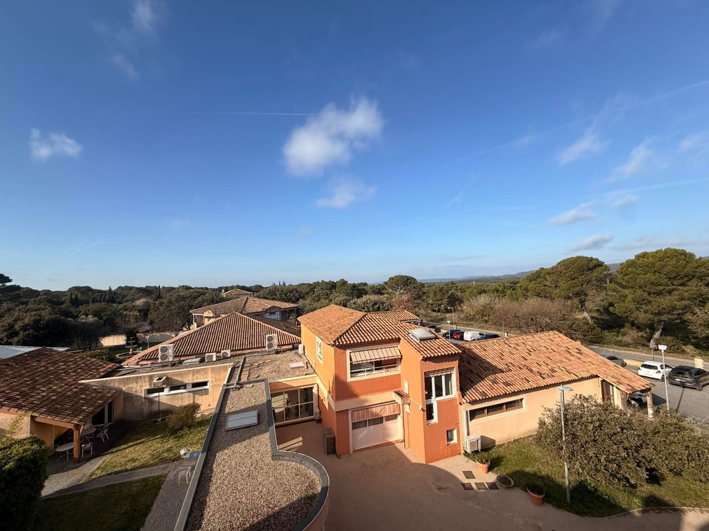 Vue ensoleillée de maisons aux tuiles orange et d'arbres sous un ciel bleu parsemé de nuages