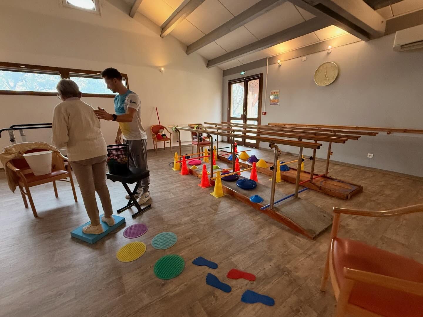 Salle de thérapie intérieure avec matériel d'équilibre, cônes, repères au sol et deux personnes debout près d'une table.